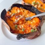 A baked sweet potato split open and topped with herbs and chopped nuts, served on a white plate. A fork is inserted into the soft, orange flesh of these crispy roasted sweet potatoes.