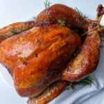 An Easy Thanksgiving Turkey, golden-brown and roasted to perfection, sits on a white plate adorned with fresh herbs atop a white napkin, against a plain white background.