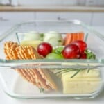 A glass container filled with sliced crackers, cheese, green and red grapes, carrot sticks, and cucumber slices creates the perfect Adult Lunchable, placed on a white surface with a blurred kitchen background.