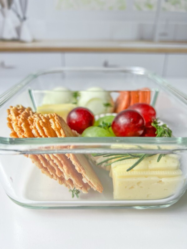 A glass container filled with sliced crackers, cheese, green and red grapes, carrot sticks, and cucumber slices creates the perfect Adult Lunchable, placed on a white surface with a blurred kitchen background.