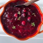 A bright orange bowl filled with thick cranberry sauce, garnished with whole cranberries and small green herb sprigs, showcases a classic Cranberry Sauce Recipe, with a wooden spoon resting in the bowl.