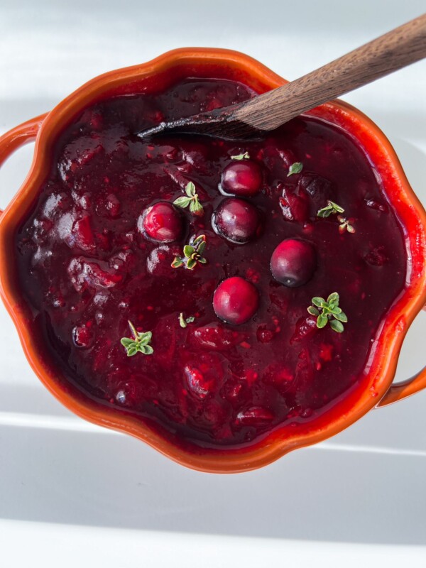 A bright orange bowl filled with thick cranberry sauce, garnished with whole cranberries and small green herb sprigs, showcases a classic Cranberry Sauce Recipe, with a wooden spoon resting in the bowl.