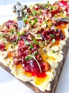 A close-up of a wooden serving board styled as a Holiday Butter Board, topped with whipped cheese, assorted fruit jams, chopped nuts, fresh herbs, and a decorative cheese knife.