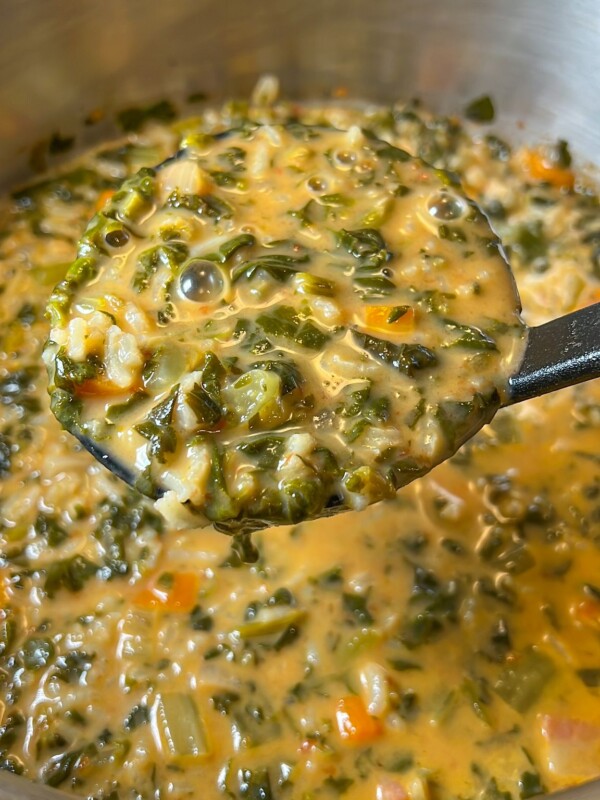 A close-up of a creamy spinach soup recipe with chopped greens, carrots, and rice, being scooped from a stainless steel pot with a black ladle.