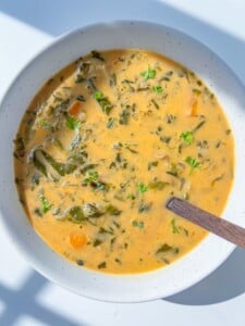 A bowl of creamy spinach soup with leafy greens, carrots, and herbs, served in a white bowl with a wooden spoon on a sunlit surface—an inviting Spinach Soup Recipe to savor.