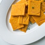 Rectangular, orange-colored crackers or chips stacked on a white textured plate, with a few pieces garnished with small green herbs—these snacks are inspired by the savory flavor of Vegetable Stock Cubes.