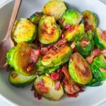 A white bowl filled with Bacon Brussels Sprouts and crispy bacon pieces, with a wooden spoon resting on the side.