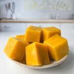 A small plate holds several bright yellow-orange frozen cubes, possibly Homemade Chicken Bouillon Cubes or fruit puree, set on a white kitchen counter with a blurred background.