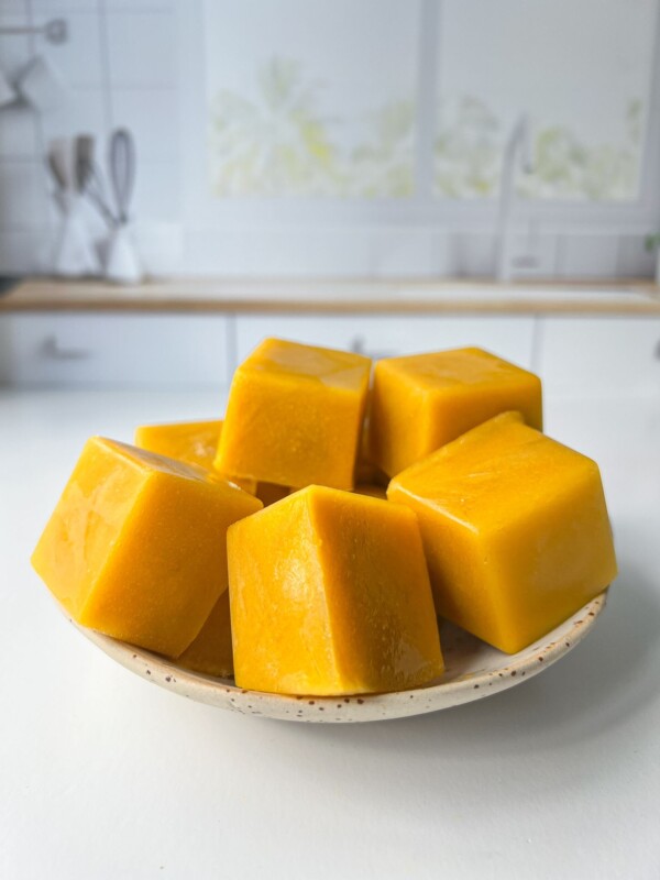 A small plate holds several bright yellow-orange frozen cubes, possibly Homemade Chicken Bouillon Cubes or fruit puree, set on a white kitchen counter with a blurred background.