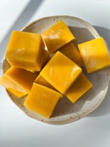 A small ceramic plate holds several bright yellow, glossy homemade chicken bouillon cubes, arranged in a loose pile on a white surface with sunlight casting soft shadows.