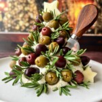 A festive Christmas Tree Cheese Ball appetizer, made of herbed cheese, green and black olives, and rosemary sprigs, sits on a white plate with a wooden-handled cheese knife and star-shaped cheese pieces.