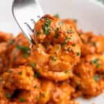 A close-up of a fork holding a piece of cooked shrimp in a spicy red sauce, garnished with herbs. More shrimp covered in the same sauce are visible on a white plate in the background.
