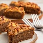 Close-up of a slice of walnut-topped cake on a plate. The cake is moist and brown, with a nutty topping. In the background, more slices are visible on plates, and two forks are placed beside them. The setting is on a light-colored surface.