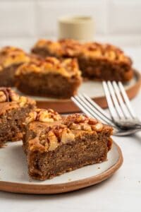 Close-up of a slice of walnut-topped cake on a plate. The cake is moist and brown, with a nutty topping. In the background, more slices are visible on plates, and two forks are placed beside them. The setting is on a light-colored surface.