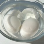 Two peeled hard-boiled eggs sit in a clear glass bowl filled with water, bits of eggshell floating around them. Bathed in natural sunlight, they’re ready to be sliced for delicious egg sandwiches.