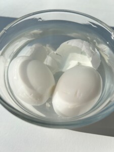Two peeled hard-boiled eggs sit in a clear glass bowl filled with water, bits of eggshell floating around them. Bathed in natural sunlight, they’re ready to be sliced for delicious egg sandwiches.