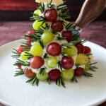 A festive Grape Christmas Tree Cheese Ball appetizer features green and red grapes, rosemary sprigs, and cheese, topped with a star-shaped cheese piece, arranged on a white plate with a knife inserted at the top.