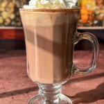 A glass mug of homemade hot chocolate topped with whipped cream and gold sprinkles sits on a table, with a warm, blurry background of yellow and red lights.