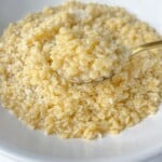 A close-up of a white bowl filled with creamy, cooked orzo pasta—also known as pastina—topped with grated cheese, with a spoon lifting a portion.