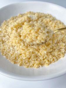 A close-up of a white bowl filled with creamy, cooked orzo pasta—also known as pastina—topped with grated cheese, with a spoon lifting a portion.