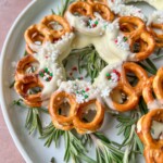 A white plate with rosemary sprigs displays a festive Pretzel Wreath, featuring pretzels dipped in white chocolate and decorated with red, green, and white sprinkles to resemble a holiday wreath.