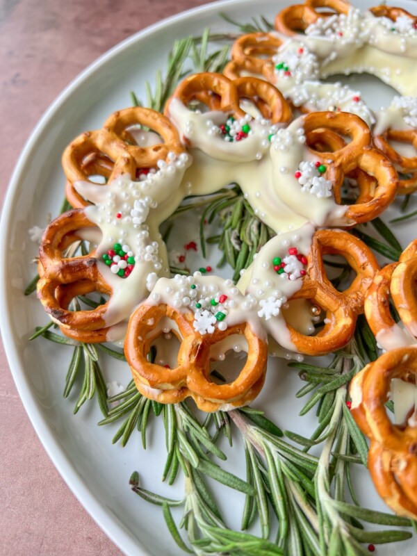 A white plate with rosemary sprigs displays a festive Pretzel Wreath, featuring pretzels dipped in white chocolate and decorated with red, green, and white sprinkles to resemble a holiday wreath.