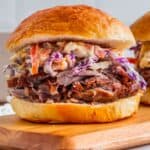 Pulled pork sandwich with coleslaw on a wooden board. The sandwich features tender, shredded pork topped with creamy coleslaw in a soft, golden bun.