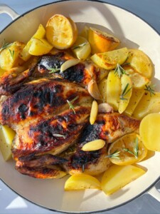 Spatchcock Greek Lemon Chicken with crispy, browned skin is served alongside golden potato wedges, lemon halves, garlic cloves, and sprigs of rosemary in a white dish.