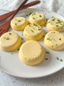 A white plate holds six creamy yellow Copycat Starbucks Egg-Bites with Ricotta, garnished with chopped herbs. Two wooden serving utensils rest on the plate’s edge against a light and airy background.
