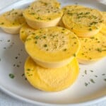 A white plate with several round, yellow Sun Dried Tomato Pesto Eggs bites sprinkled with chopped herbs, arranged in a small pile on a light surface.
