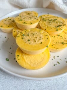 A white plate with several round, yellow Sun Dried Tomato Pesto Eggs bites sprinkled with chopped herbs, arranged in a small pile on a light surface.