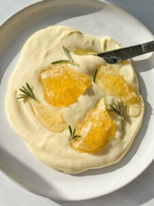 A white plate with creamy whipped Brie, topped with three pieces of honeycomb and sprigs of rosemary. A butter knife rests on the plate. The scene is brightly lit.