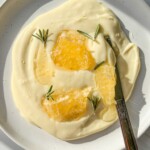 A white plate with creamy yogurt and whipped Brie, topped with honeycomb pieces and rosemary sprigs; a butter knife rests on the plate.