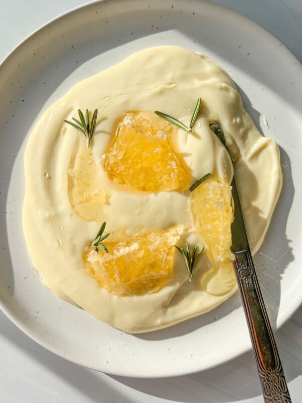 A white plate with creamy yogurt and whipped Brie, topped with honeycomb pieces and rosemary sprigs; a butter knife rests on the plate.