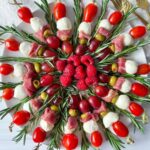 A round platter forms an Antipasti Christmas Wreath, with skewers of mozzarella balls, cherry tomatoes, grapes, olives, prosciutto, and rosemary arranged in a circle; raspberries fill the center and two gold forks rest on the side.