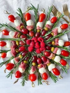 A round platter forms an Antipasti Christmas Wreath, with skewers of mozzarella balls, cherry tomatoes, grapes, olives, prosciutto, and rosemary arranged in a circle; raspberries fill the center and two gold forks rest on the side.