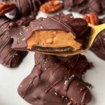A plate of Healthy Chocolate Turtles—pecan caramel clusters dipped in rich chocolate, with one on a gold fork revealing gooey caramel and pecans, finished with a sprinkle of sea salt.