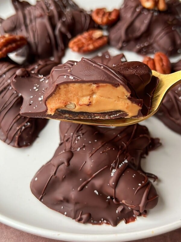 A plate of Healthy Chocolate Turtles—pecan caramel clusters dipped in rich chocolate, with one on a gold fork revealing gooey caramel and pecans, finished with a sprinkle of sea salt.