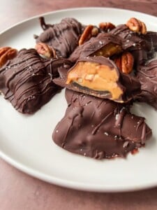 A close-up of Healthy Chocolate Turtles—chocolate-covered candies with a caramel and nut filling, topped with pecans and a sprinkle of sea salt, beautifully arranged on a white plate.