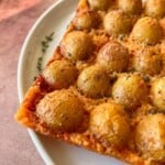 A close-up of a rectangular slice of pizza on a white plate, topped with crispy Parmesan potatoes and herbs, featuring a golden-brown crust.