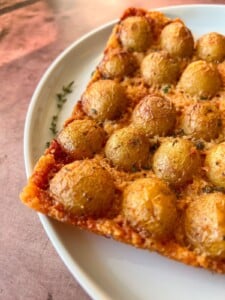 A close-up of a rectangular slice of pizza on a white plate, topped with crispy Parmesan potatoes and herbs, featuring a golden-brown crust.