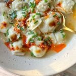 A bowl of Dumpling Soup in broth, topped with chili oil, sesame seeds, chopped chives, and drizzled sauce, served with a golden spoon on the side.