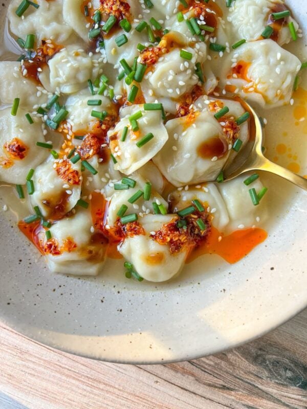 A bowl of Dumpling Soup in broth, topped with chili oil, sesame seeds, chopped chives, and drizzled sauce, served with a golden spoon on the side.