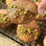 A hand holds a chocolate-dipped honey cookie sprinkled with chopped pistachios. More honey cookies rest on a wire cooling rack in the background.