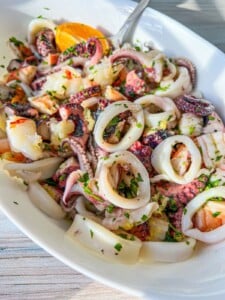 A white dish filled with a vibrant Seafood Salad featuring sliced calamari rings, shrimp, and octopus tentacles, garnished with chopped parsley and an orange wedge. A fork rests in the dish.