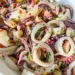 Close-up of a seafood salad featuring rings and tentacles of octopus, garnished with chopped parsley and garlic, served in a white bowl.