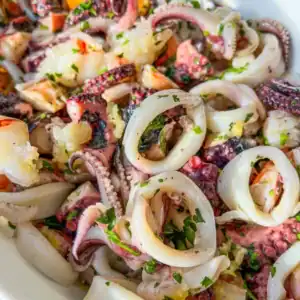 Close-up of a seafood salad featuring rings and tentacles of octopus, garnished with chopped parsley and garlic, served in a white bowl.