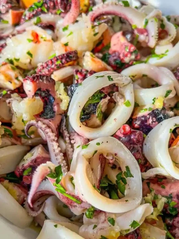 Close-up of a seafood salad featuring rings and tentacles of octopus, garnished with chopped parsley and garlic, served in a white bowl.