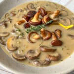A comforting Cream of Mushroom Soup topped with sautéed mushrooms and a sprig of fresh thyme, served in a speckled bowl with a yellow spoon.
