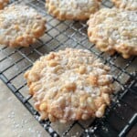Close-up of classic Pignoli Cookies topped with pine nuts and dusted with powdered sugar, cooling on a black wire rack. More cookies and scattered sugar can be seen in the background.
